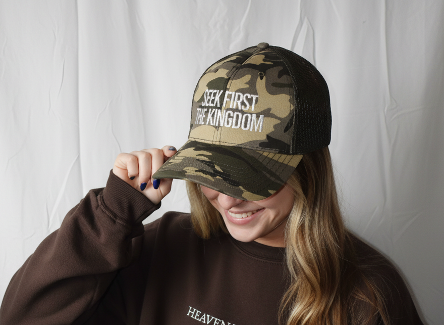 Person wearing a brown sweatshirt with text and a camouflage cap with text, against a white background
