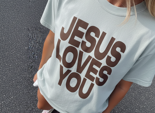 Person wearing a light blue t-shirt with 'Jesus Loves You' printed in brown on a black asphalt background.