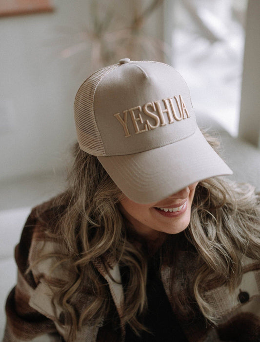 Person wearing a beige cap with 'Yeshua' embroidered on it, sitting indoors.
