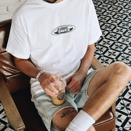 Person wearing a white t-shirt with 'Jesus Saves' text, sitting on a chair holding a coffee cup.
