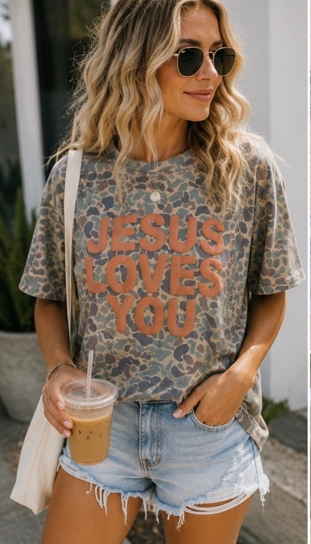 A woman wearing a distressed leopard-print graphic t-shirt with the text 'JESUS LOVES YOU' in orange, paired with light-wash denim shorts and sunglasses, holding an iced coffee.